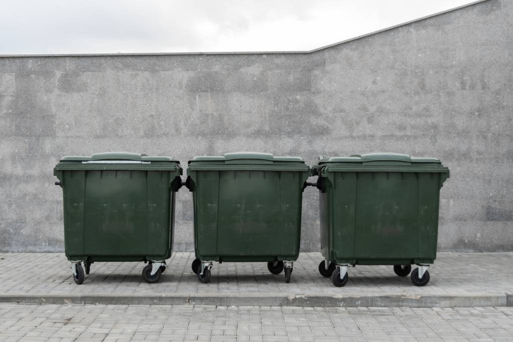 Three green large wheelie bins stand in a row against a grey wall.