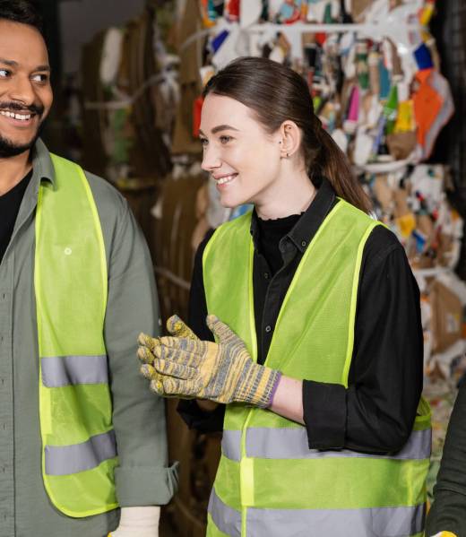 Cheerful female worker in protective vest and gloves looking away while standing near multiethnic